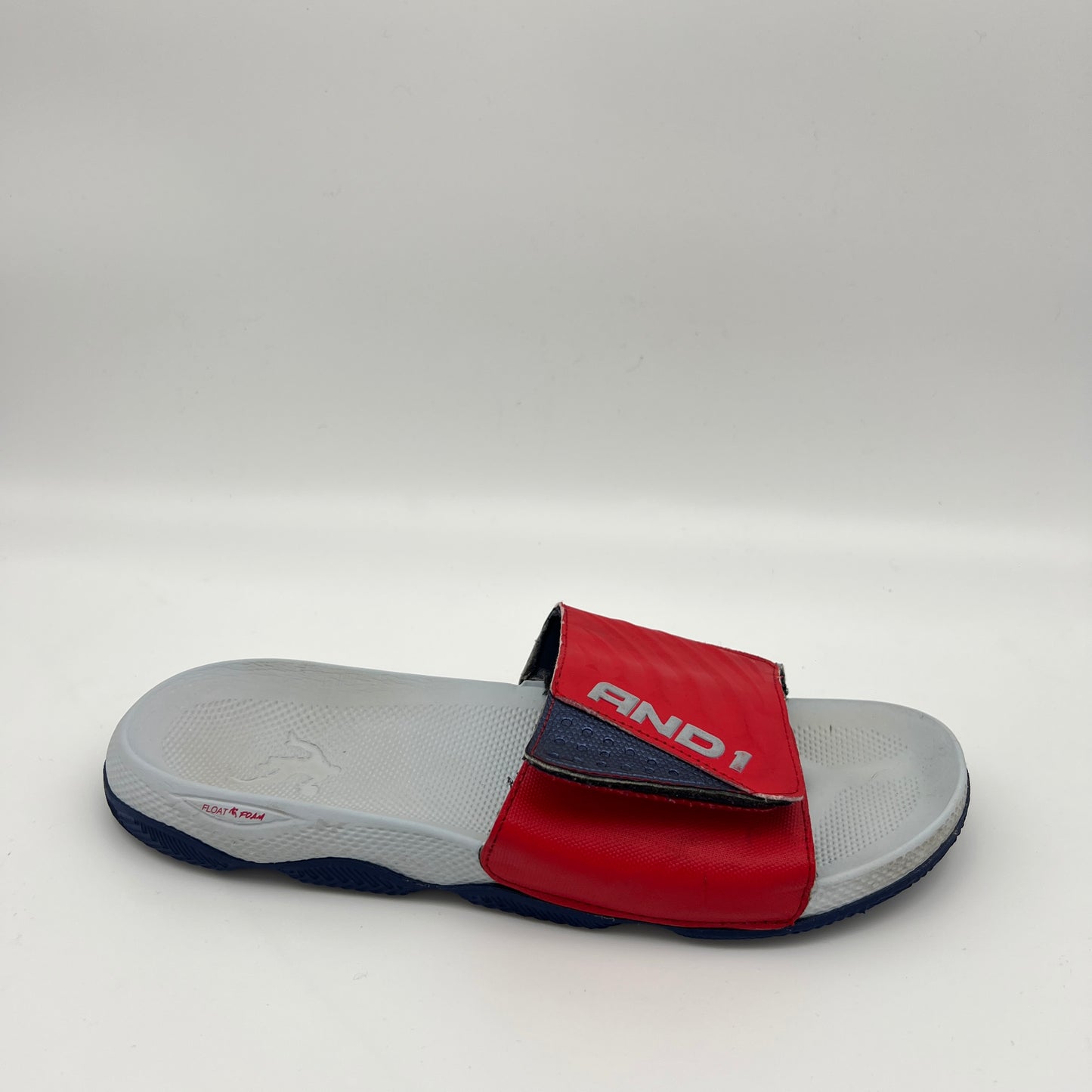 A side view of an AND1 Swish 2.0 slide sandal featuring a red adjustable strap with a navy base, metallic AND1 logo, and a contoured grey Float Foam footbed.

