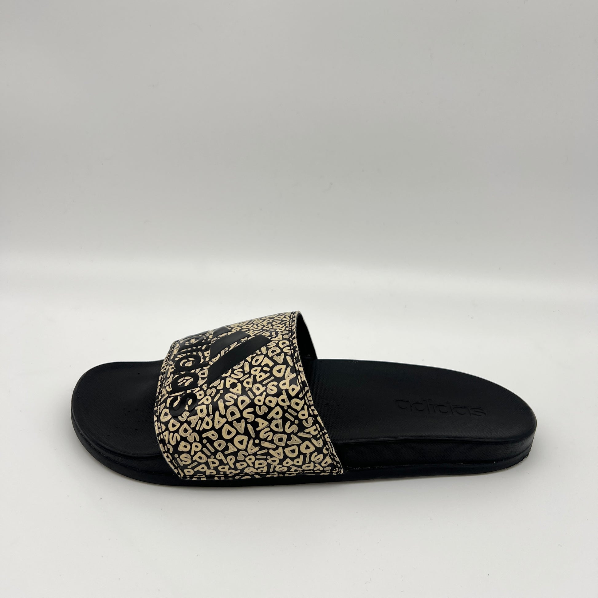 Side view of a black Adidas Adilette Comfort slide on a white background, featuring a beige and black monogram logo print strap and a plush black footbed.
