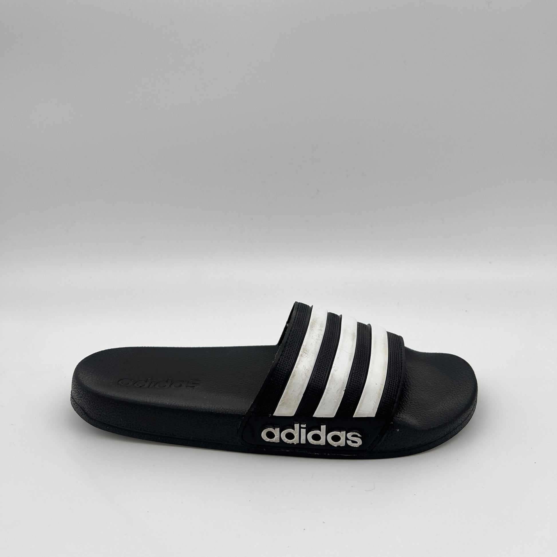 Side profile of a black Adidas Adilette Shower slide with three white stripes on the strap and a white Adidas logo on the black sole against a white background.
