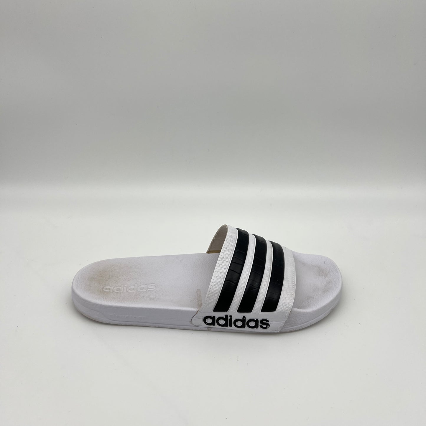 A side profile of a white Adidas Adilette Shower slide with black three-stripes on the strap and a contoured Cloudfoam footbed.