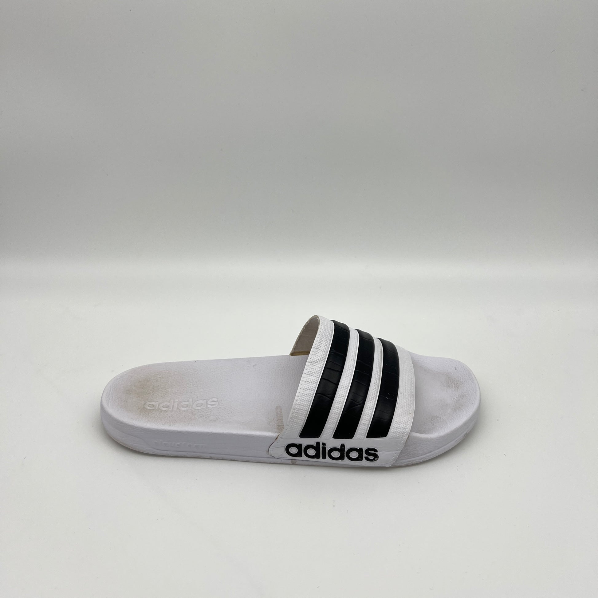 A side profile of a white Adidas Adilette Shower slide with black three-stripes on the strap and a contoured Cloudfoam footbed.