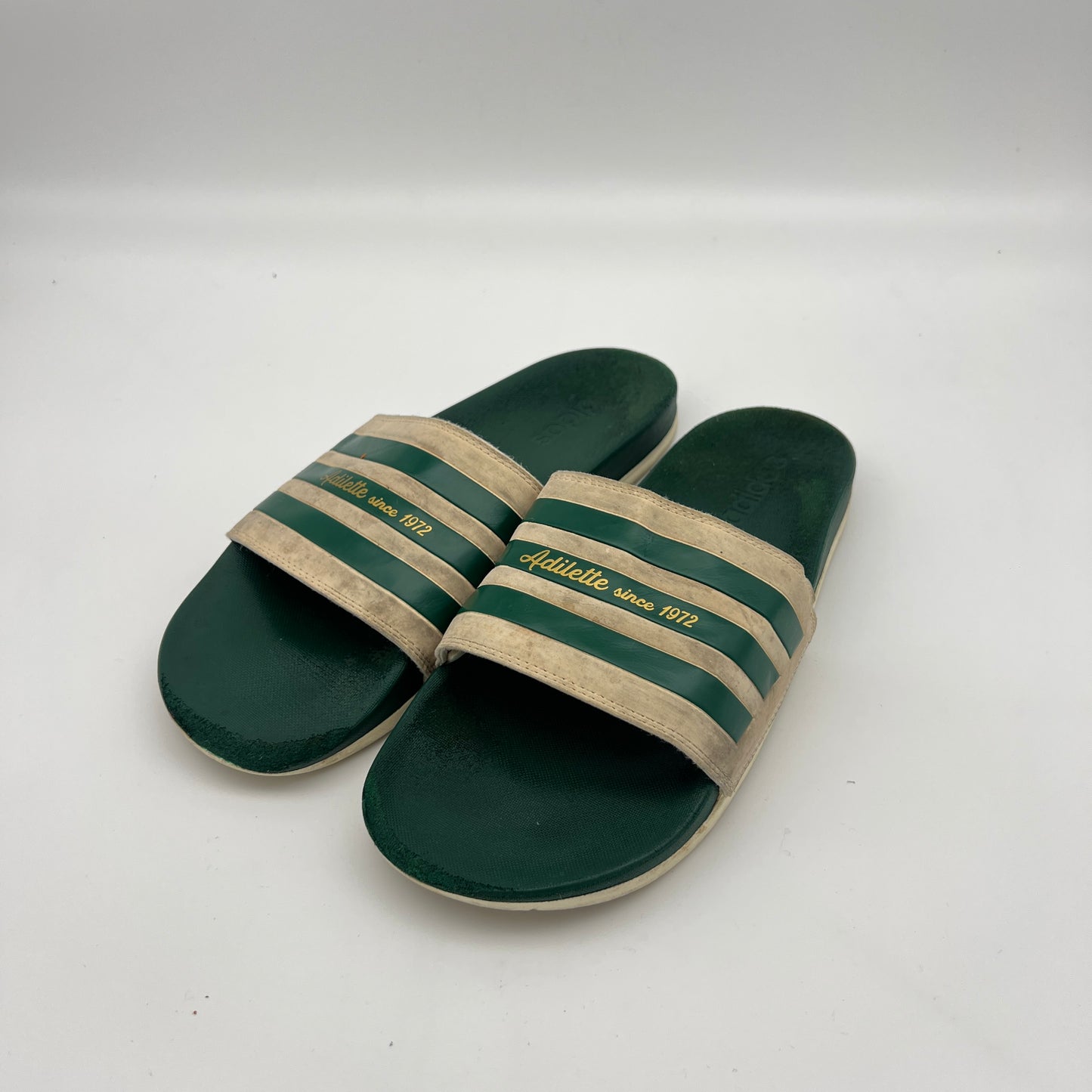 Side view of retro Adidas Adilette sandals with green 3-Stripes and gold-foil "Since 1972" lettering on an off-white strap.