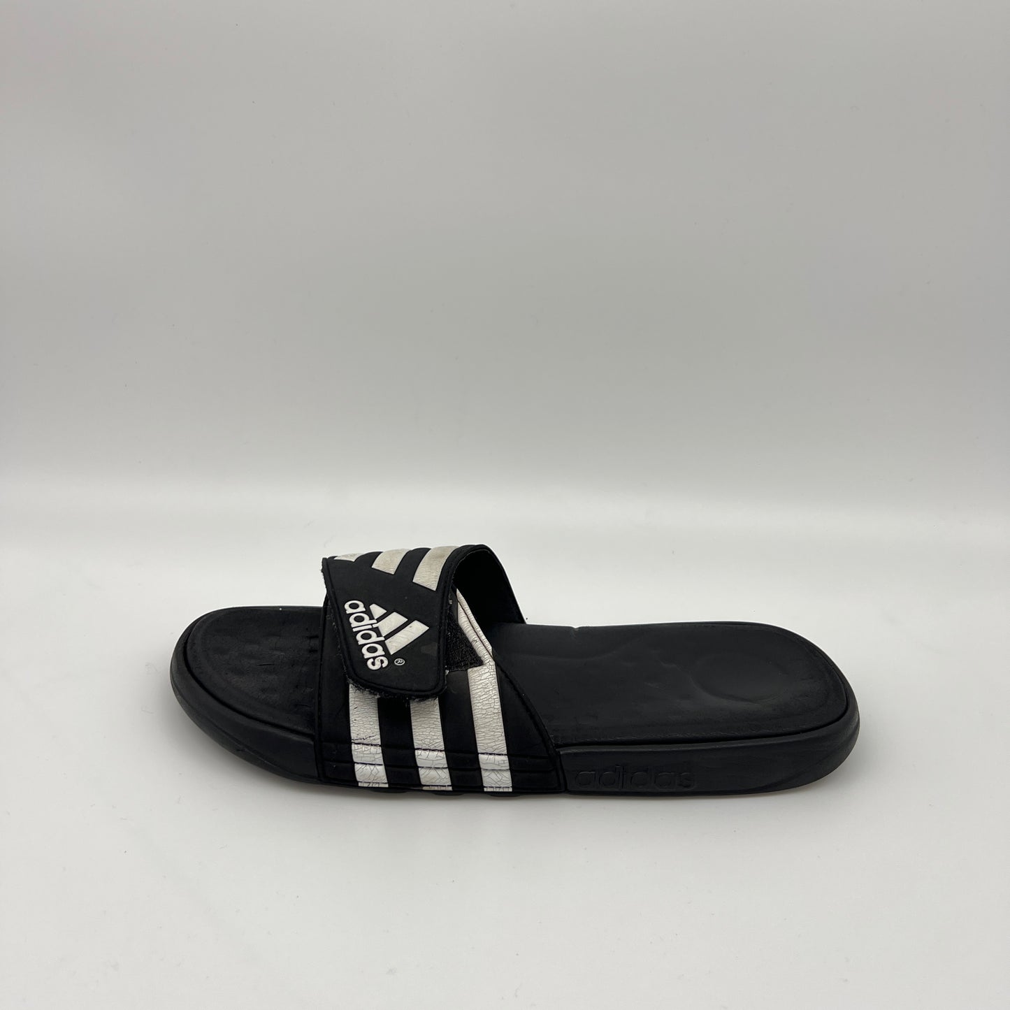 Side profile of a black Adidas Adissage Cloudfoam slide with three white stripes on an adjustable velcro strap and a smooth, cushioned black footbed.
