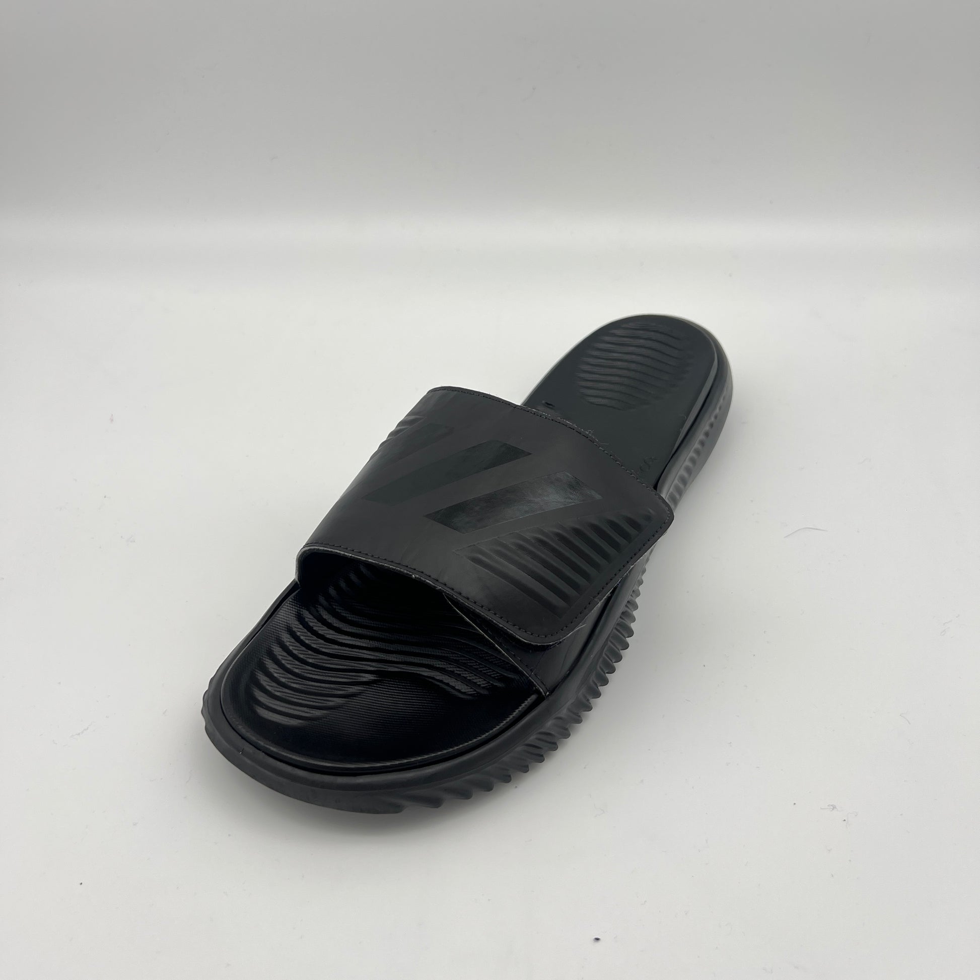 Side view of all-black Adidas Alphabounce Slides featuring a textured footbed and an adjustable velcro strap with the embossed 3-stripe logo.