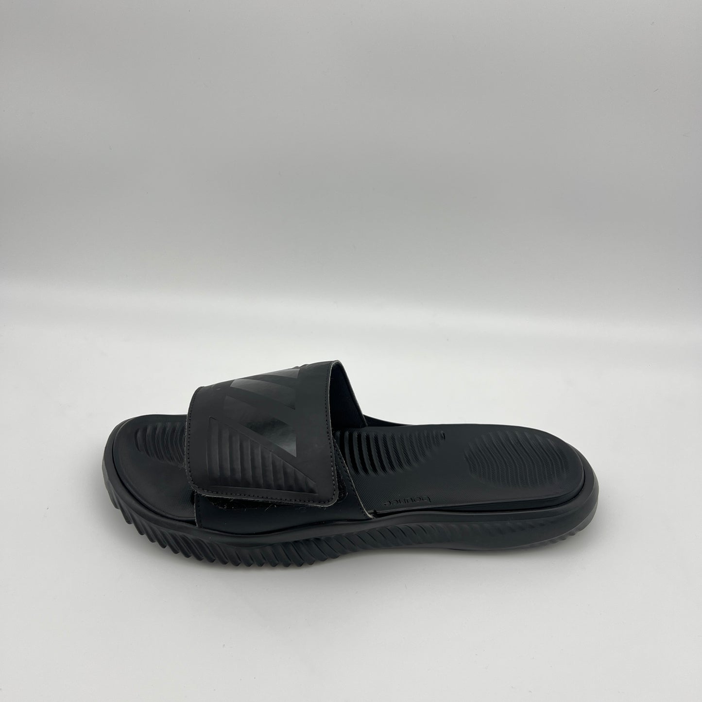 A side profile of an all-black Adidas Alphabounce slide featuring an adjustable strap with debossed branding and a thick, contoured Bounce cushioned sole.