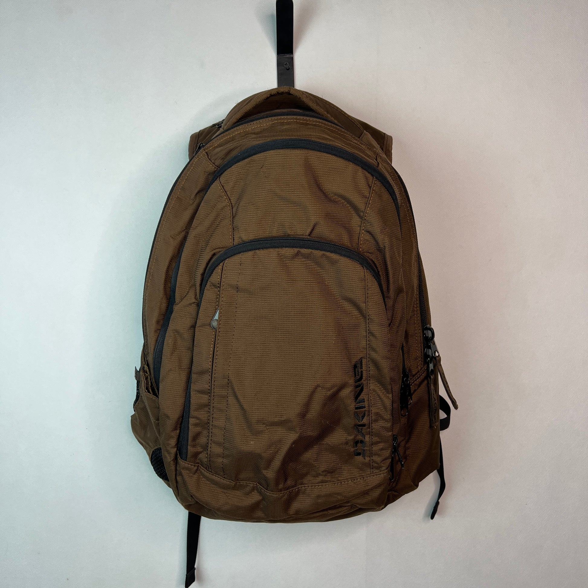 Front view of a brown textured Dakine backpack with several zippered compartments hanging from a black hook on a white wall