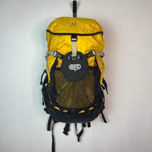 Front view of a yellow and black Haglöfs ORC 35 technical backpack with a large mesh front pocket hanging on a white wall.