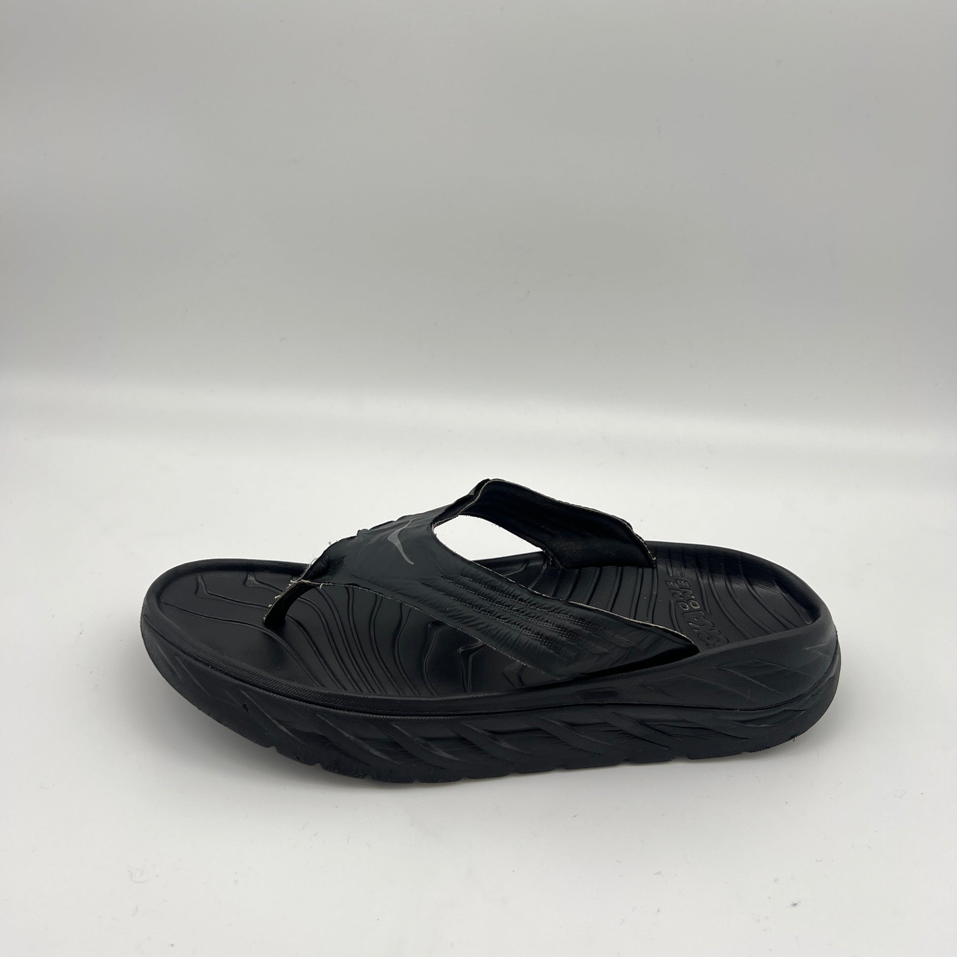 Side view of a black Hoka One One Ora Recovery Flip sandal with a thick cushioned midsole and textured footbed on a white background