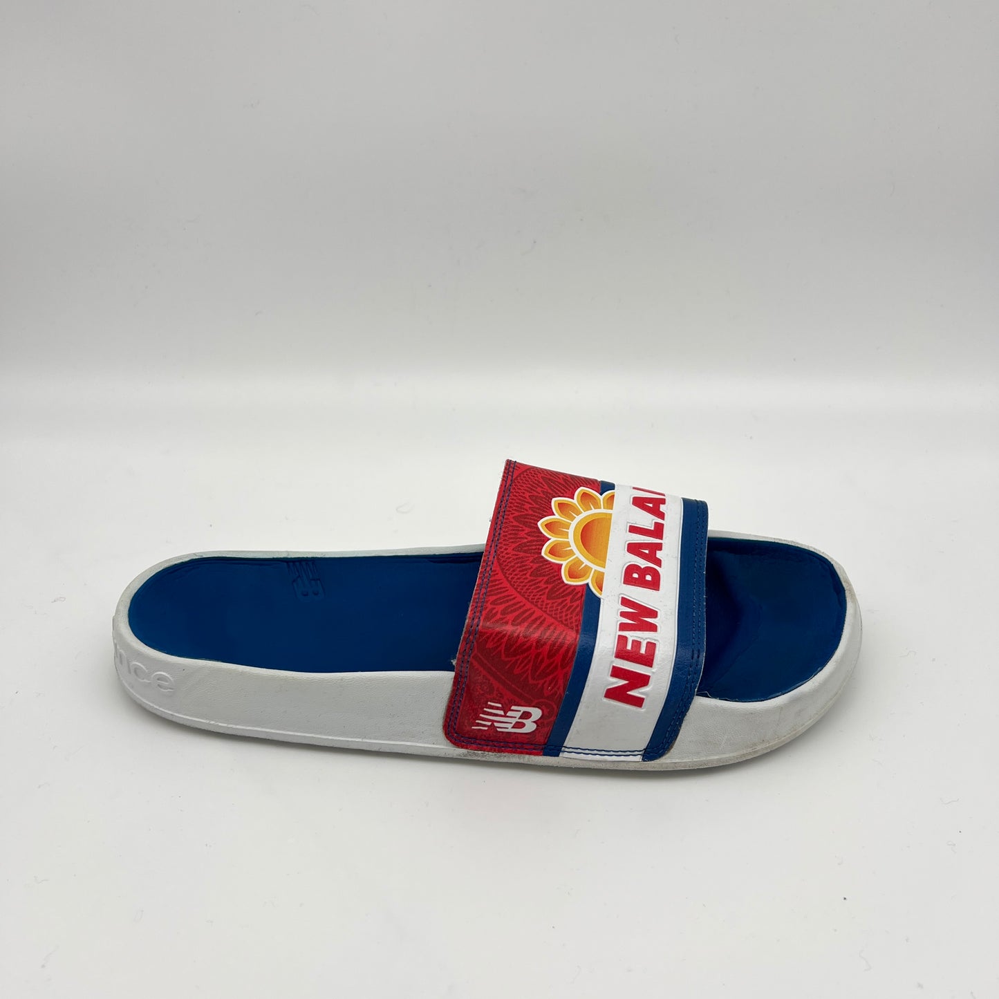 Side view of a New Balance 200 slide sandal featuring a blue footbed and a red and white strap with a yellow sunflower logo and "NEW BALANCE" in red block letters.
