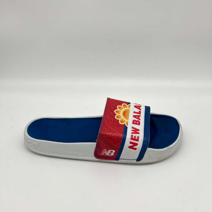 Side view of a New Balance 200 slide sandal featuring a blue footbed and a red and white strap with a yellow sunflower logo and "NEW BALANCE" in red block letters.
