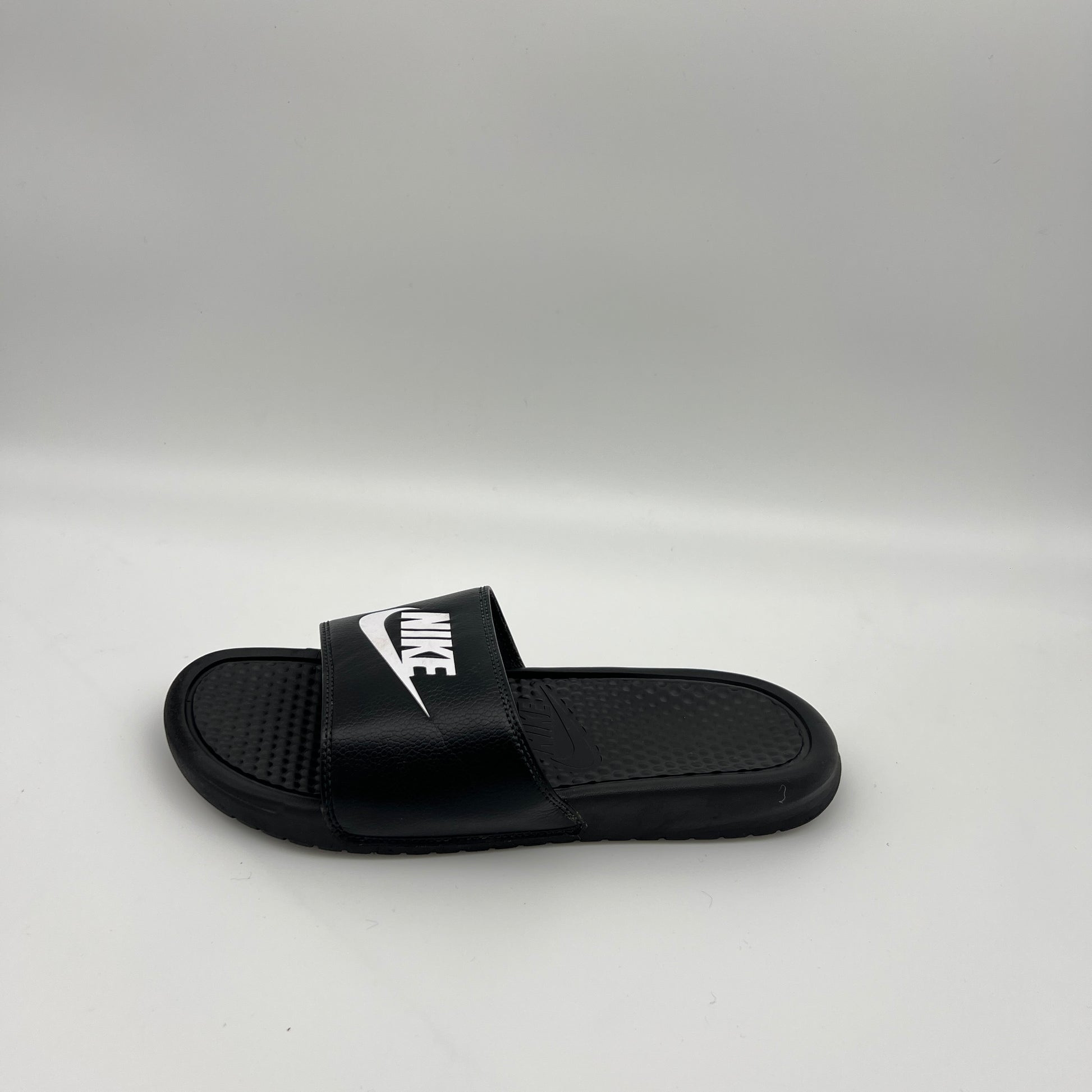 Side view of a single black Nike Benassi JDI slide sandal featuring a bold white Swoosh logo on the strap and a textured footbed on a plain white background.