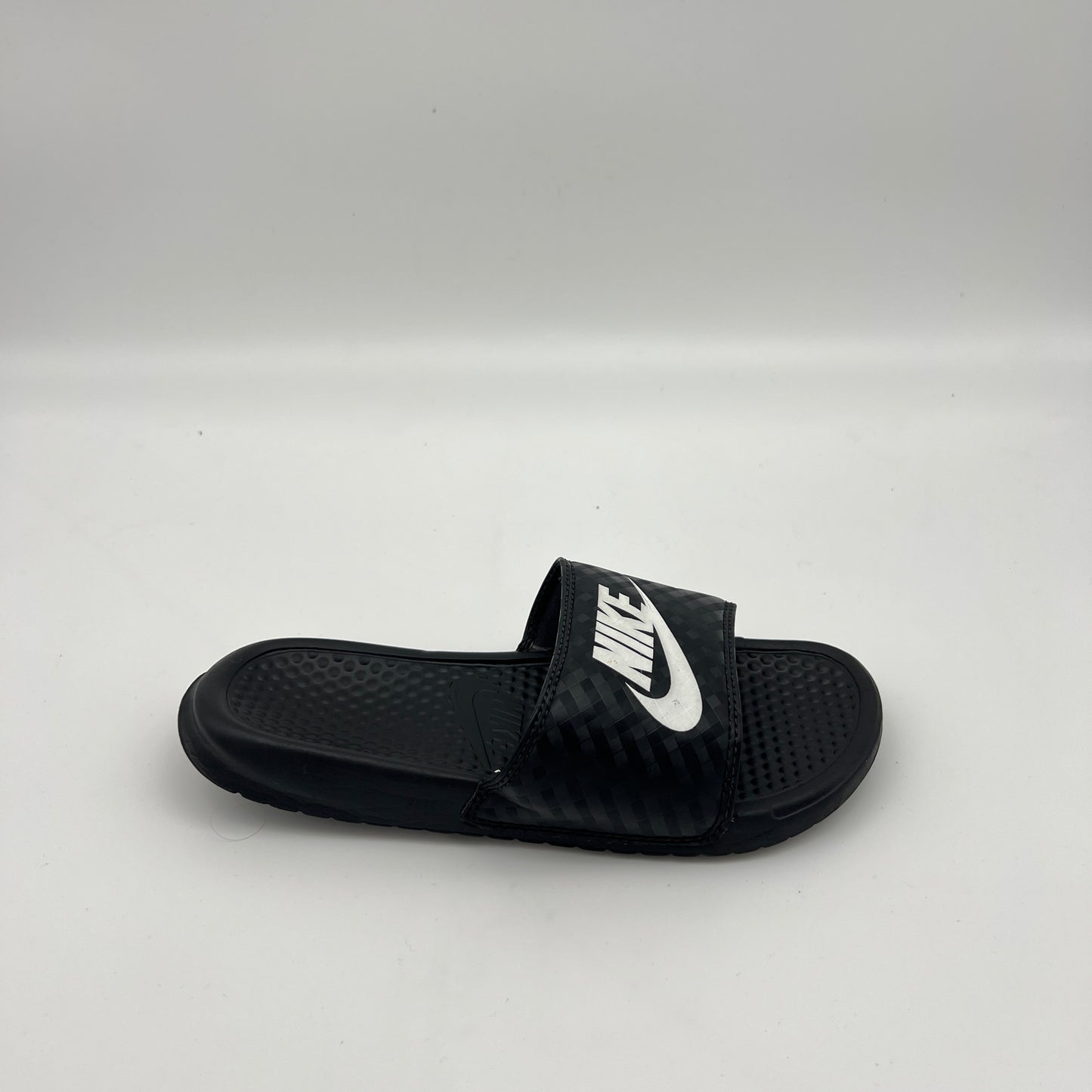 Side profile of a black Nike Benassi JDI slide sandal featuring a tonal diamond-grid pattern on the strap and a bold white Nike logo on a white background.