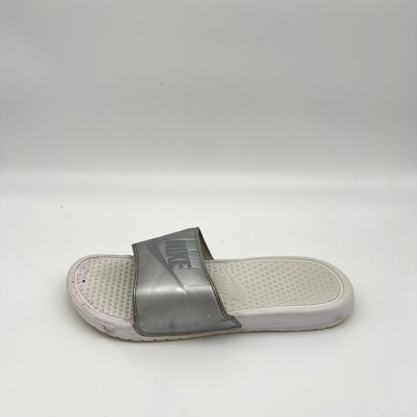 Metallic silver Nike Benassi JDI slide sandal with an embossed Nike logo on the strap and a textured white footbed, shown on a white background.