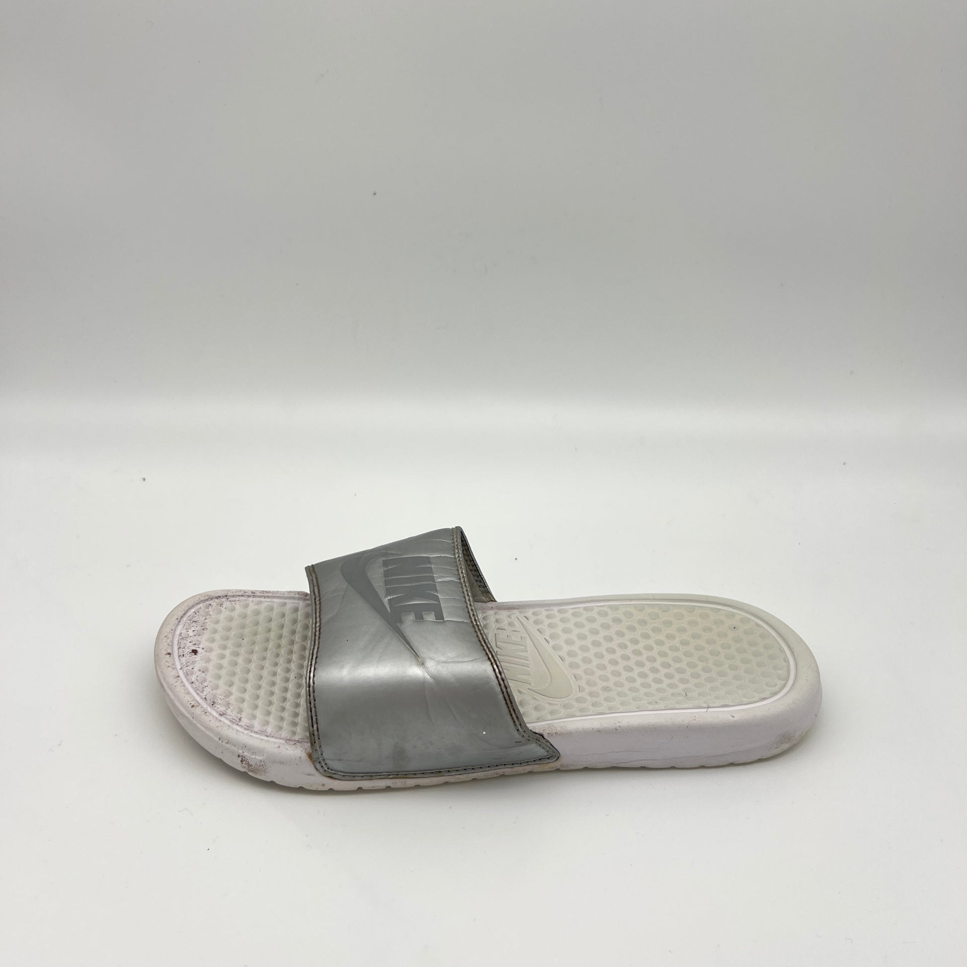Metallic silver Nike Benassi JDI slide sandal with an embossed Nike logo on the strap and a textured white footbed, shown on a white background.