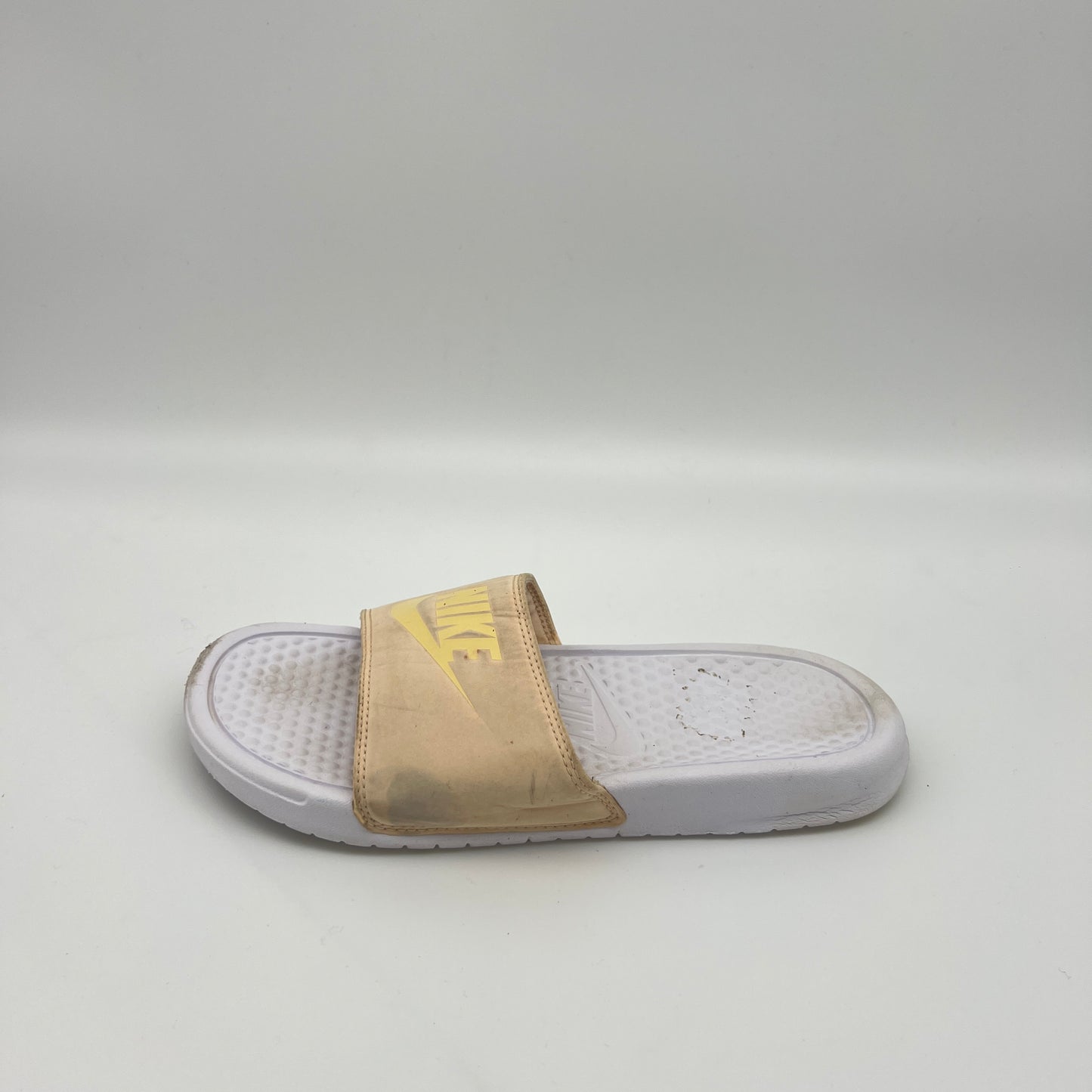 Side view of a pale ivory Nike Benassi JDI slide featuring a yellow Nike logo on the padded strap and a textured white Phylon footbed on a white background.