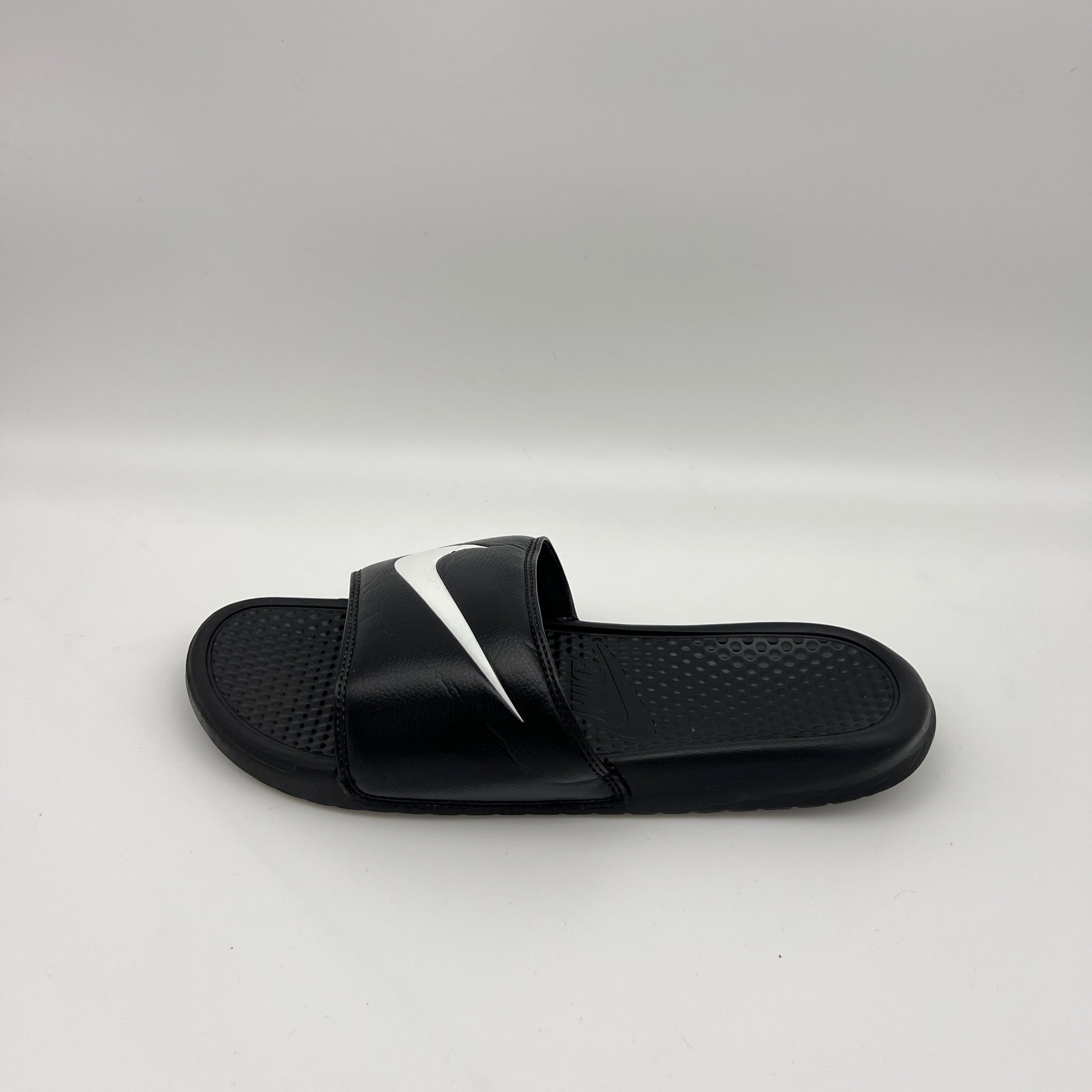 Side profile of a black Nike Benassi Swoosh slide sandal featuring a large white iconic logo on the strap and a textured footbed on a white background.