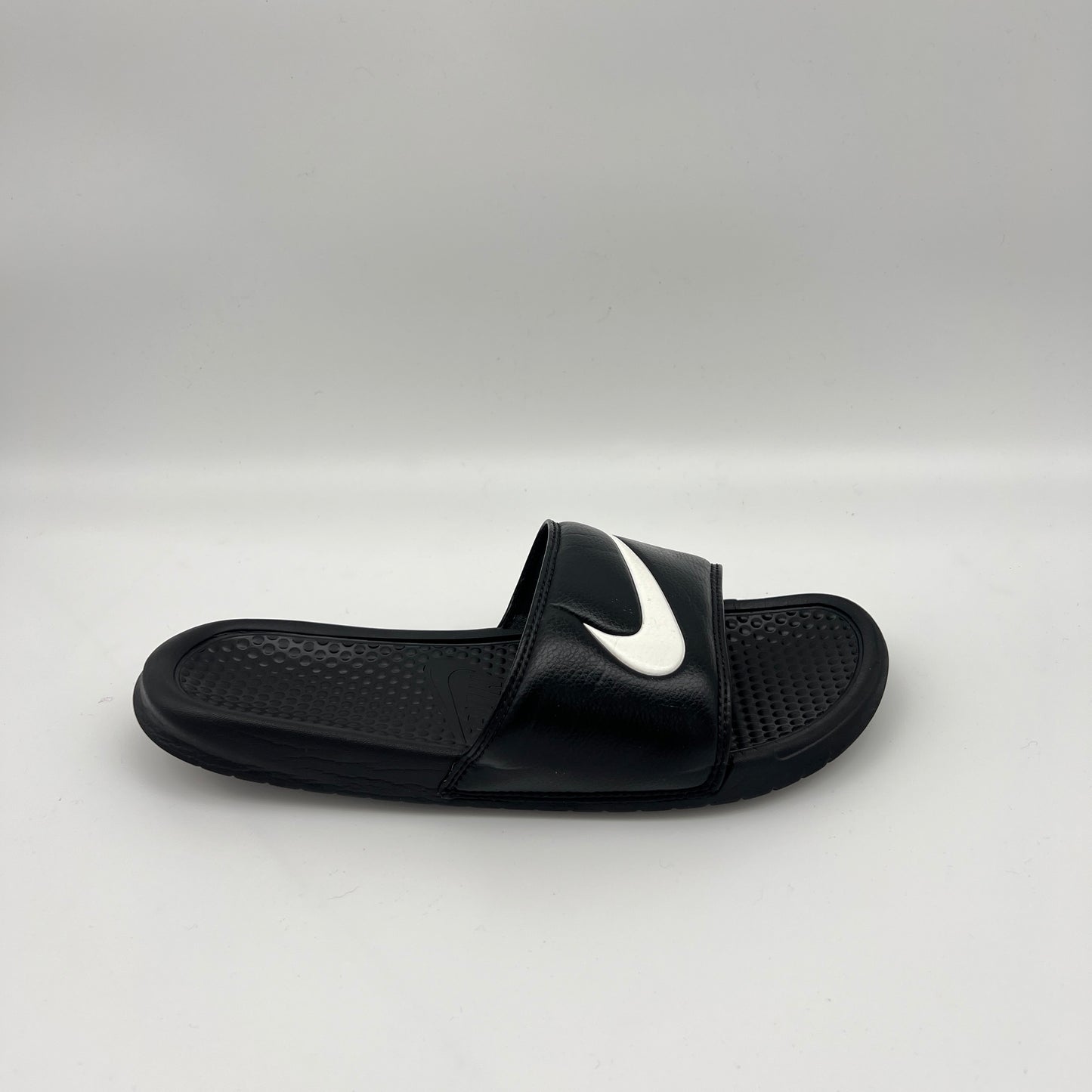 Side profile of a single black Nike Benassi Swoosh slide featuring a white Nike logo on the synthetic leather strap and a pebbled comfort footbed.
