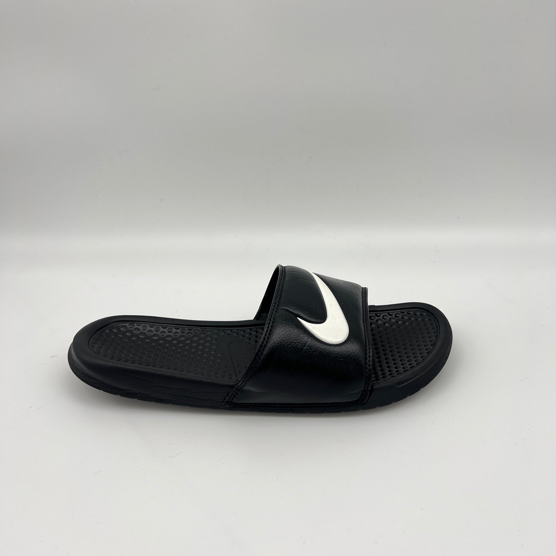 Side profile of a single black Nike Benassi Swoosh slide featuring a white Nike logo on the synthetic leather strap and a pebbled comfort footbed.
