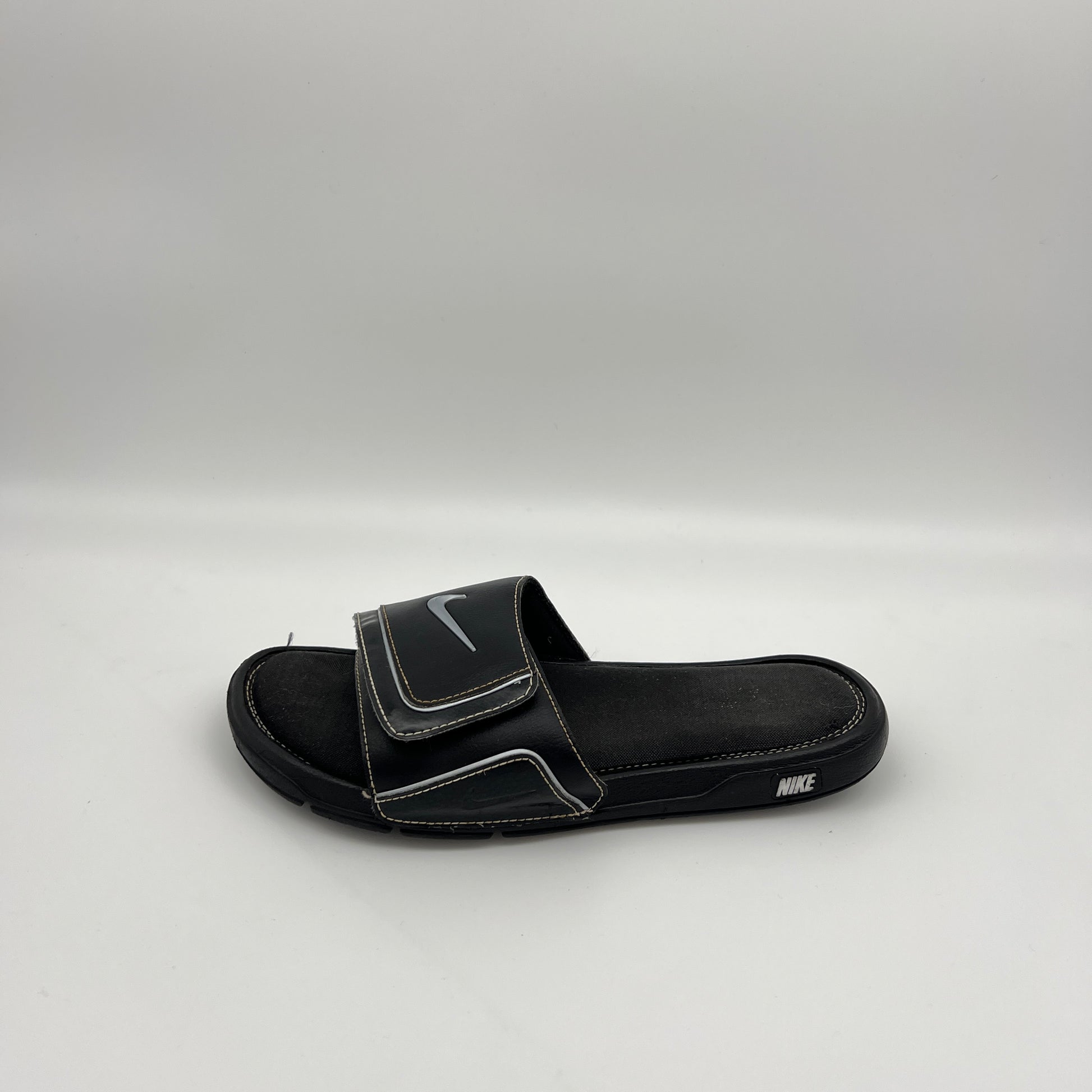 Side view of a single black Nike Comfort Slide 2 sandal with white stitching and a metallic silver Swoosh logo on an adjustable velcro strap