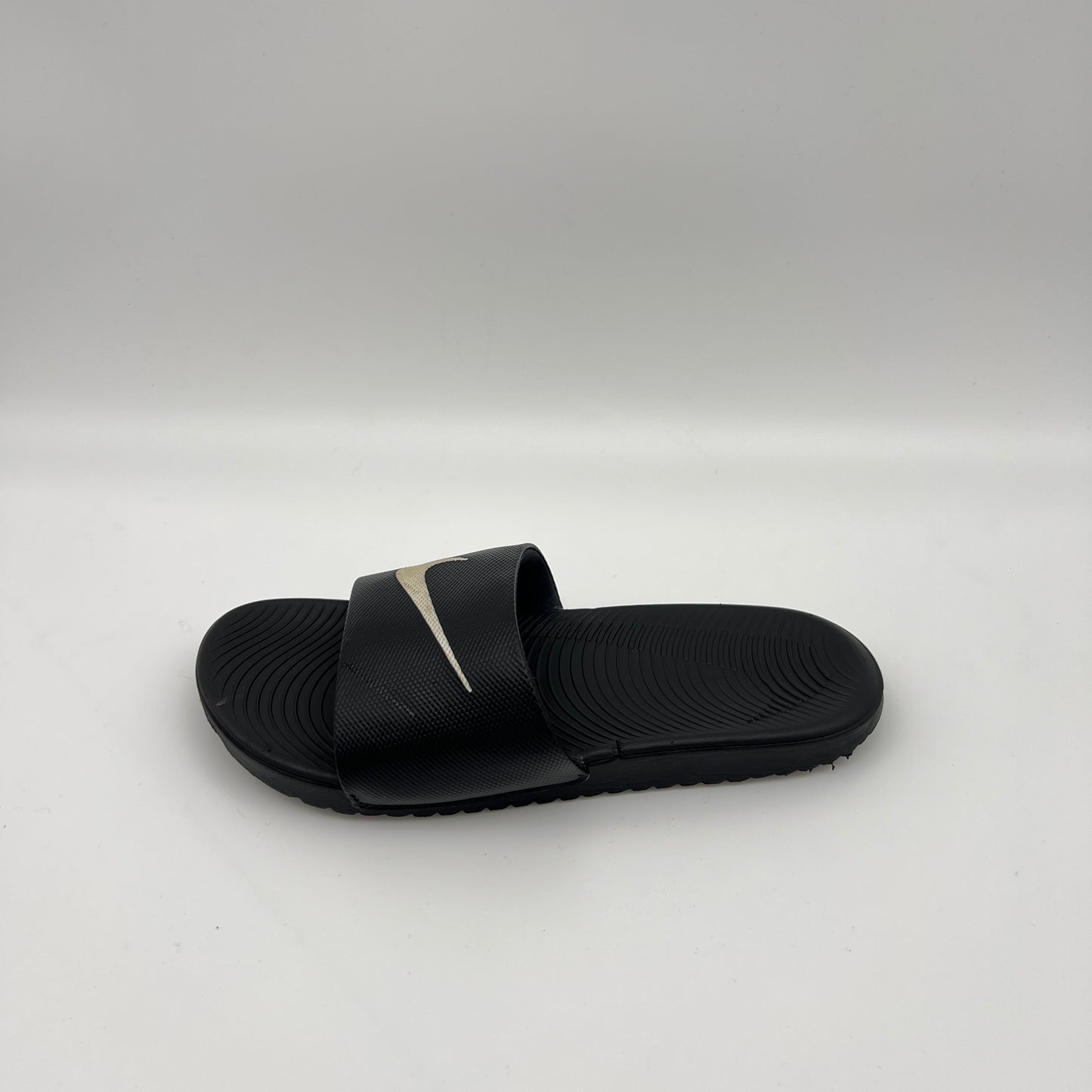 Side-angle view of a black Nike Kawa slide sandal on a white background, highlighting the metallic swoosh logo on the strap and the wavy Solarsoft footbed.