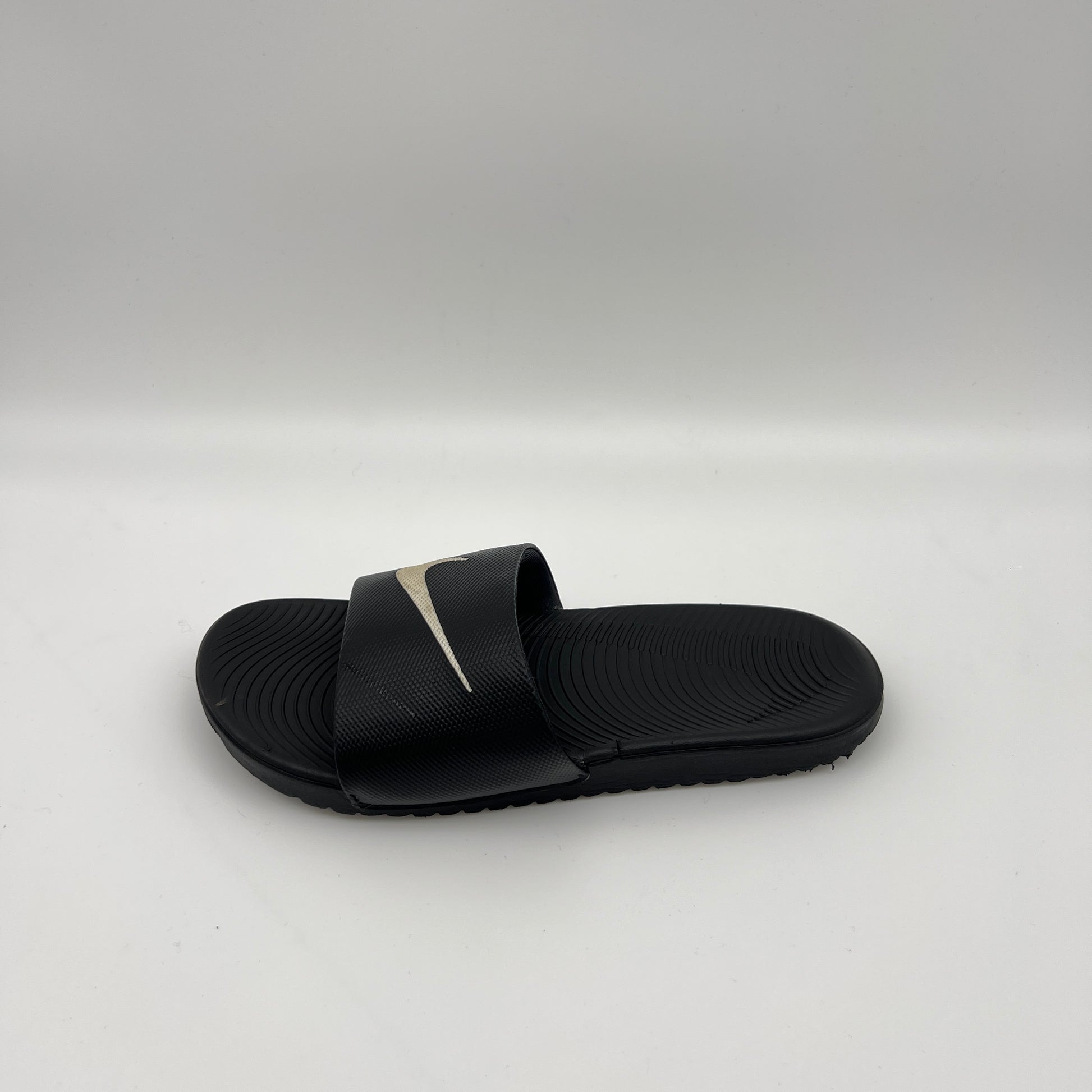Side-angle view of a black Nike Kawa slide sandal on a white background, highlighting the metallic swoosh logo on the strap and the wavy Solarsoft footbed.