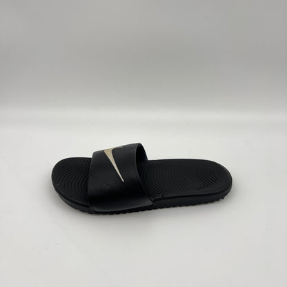 Side-angle view of a black Nike Kawa slide sandal on a white background, highlighting the metallic swoosh logo on the strap and the wavy Solarsoft footbed.