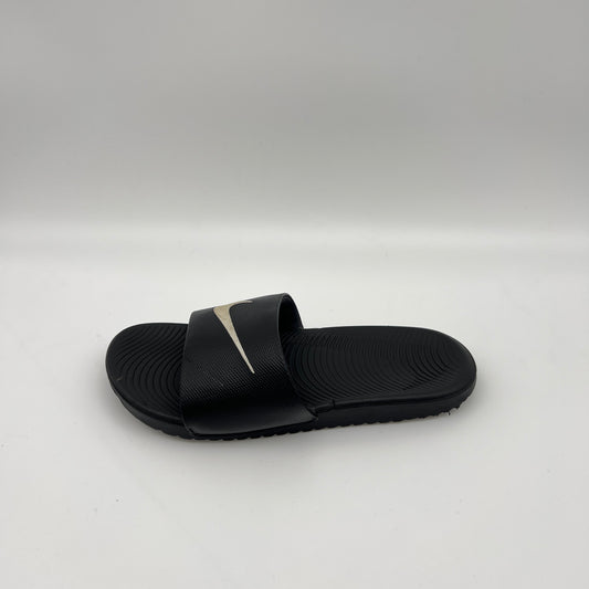 Side-angle view of a black Nike Kawa slide sandal on a white background, highlighting the metallic swoosh logo on the strap and the wavy Solarsoft footbed.