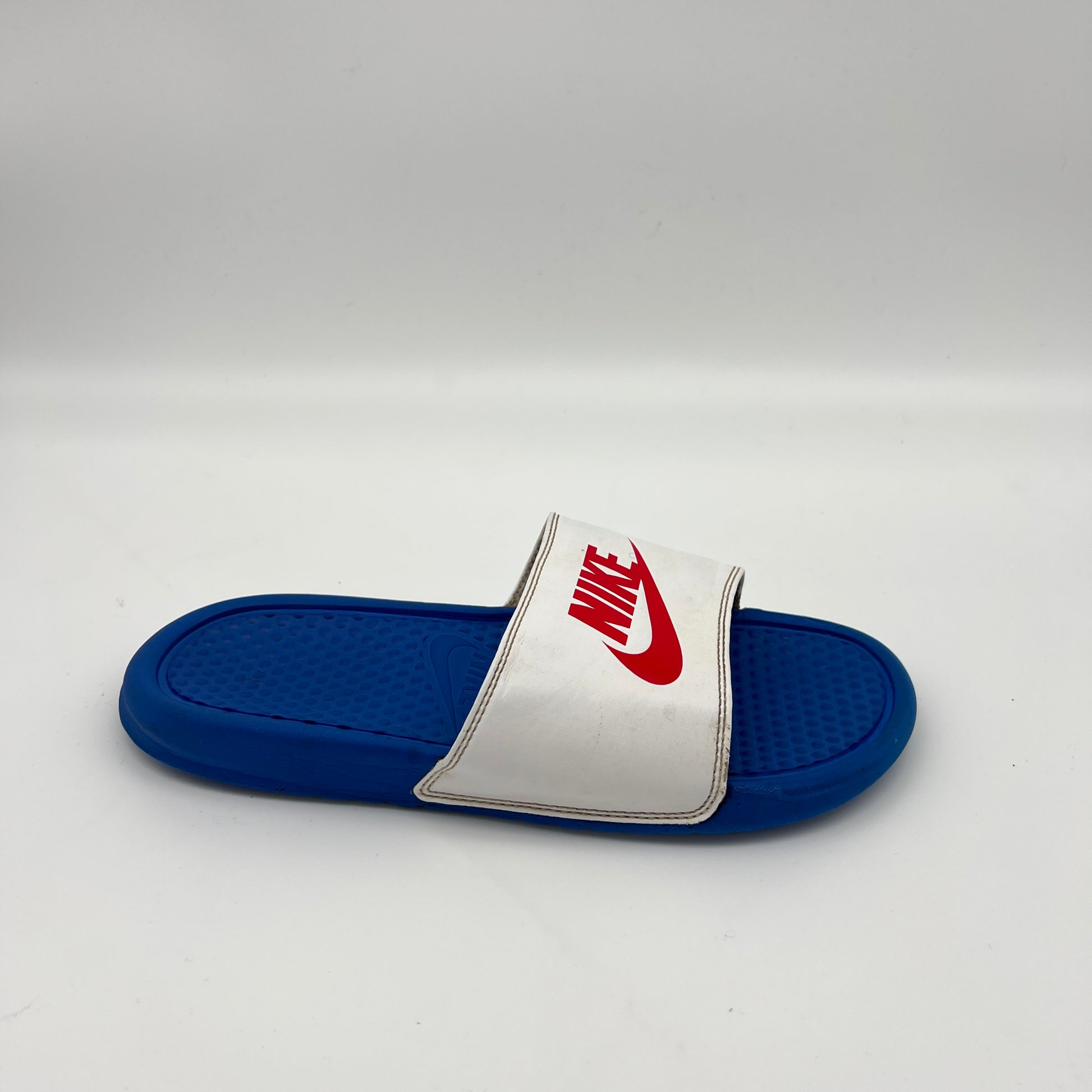 A side view of a royal blue Nike Benassi JDI slide sandal featuring a white strap with a red Nike logo and a textured blue footbed on a white background.
