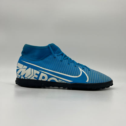 Side view of a blue Nike Mercurial Superfly 7 turf shoe featuring a white swoosh, "MERC" branding on the heel, and a black studded sole on a grey background.