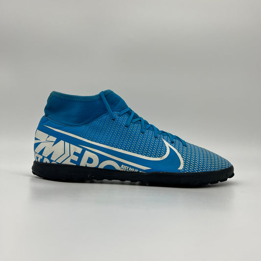Side view of a blue Nike Mercurial Superfly 7 turf shoe featuring a white swoosh, "MERC" branding on the heel, and a black studded sole on a grey background.