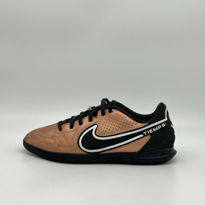 Side view of a bronze and black Nike Tiempo indoor soccer shoe with a white swoosh and black laces against a plain gray background.