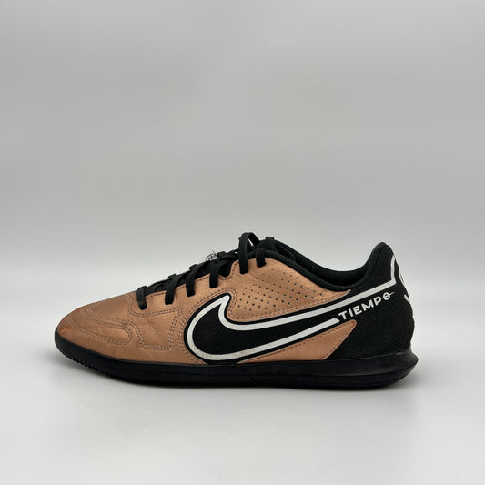 Side view of a bronze and black Nike Tiempo indoor soccer shoe with a white swoosh and black laces against a plain gray background.