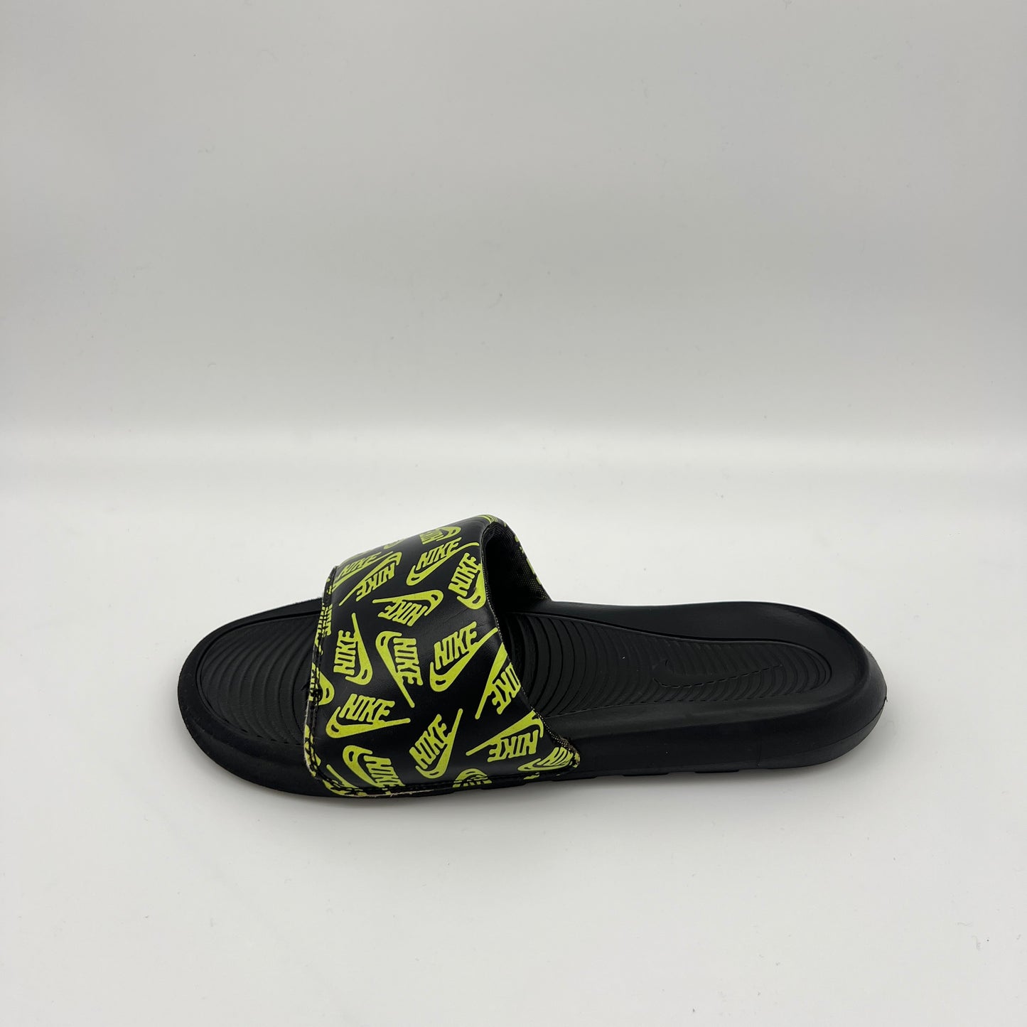 Side view of a black Nike Victori One slide sandal featuring a repeating volt green Nike logo print on the strap and a contoured black footbed.