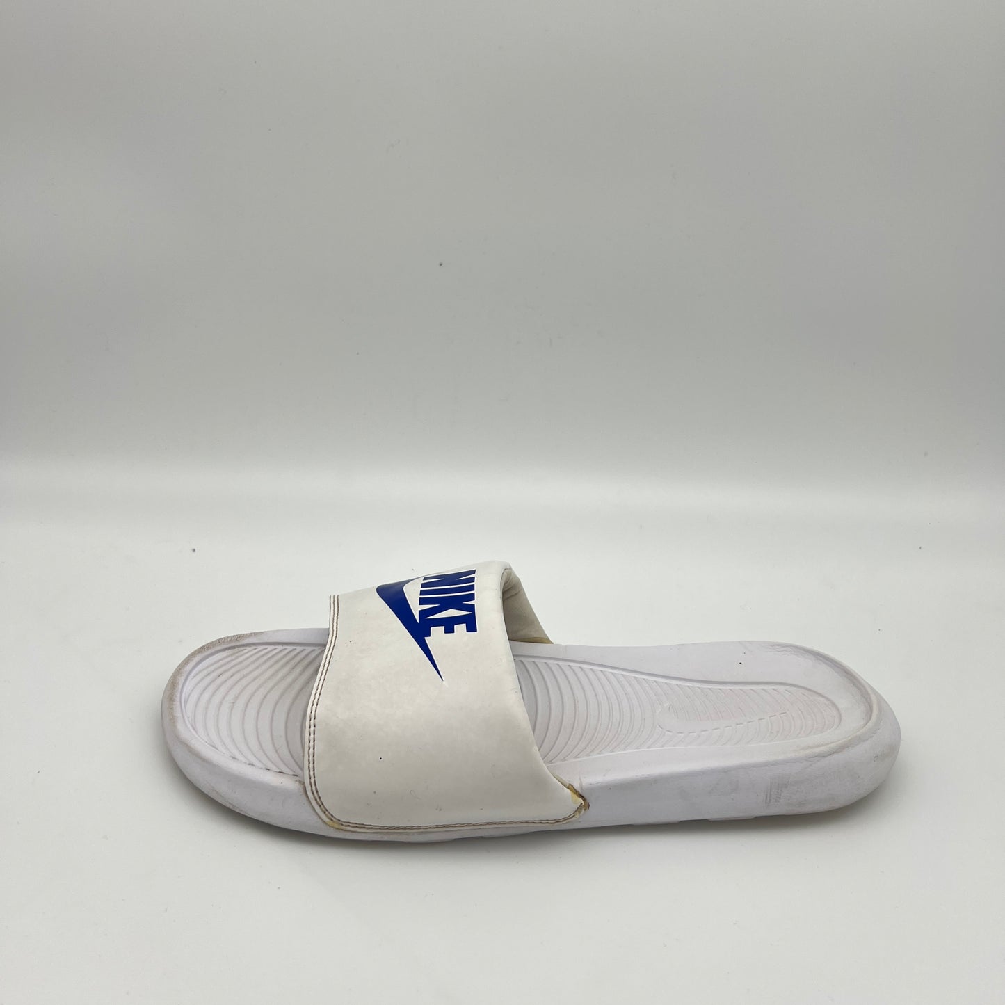 White Nike Victori One slide sandal with a royal blue Nike logo and swoosh on the padded strap, set against a white background.