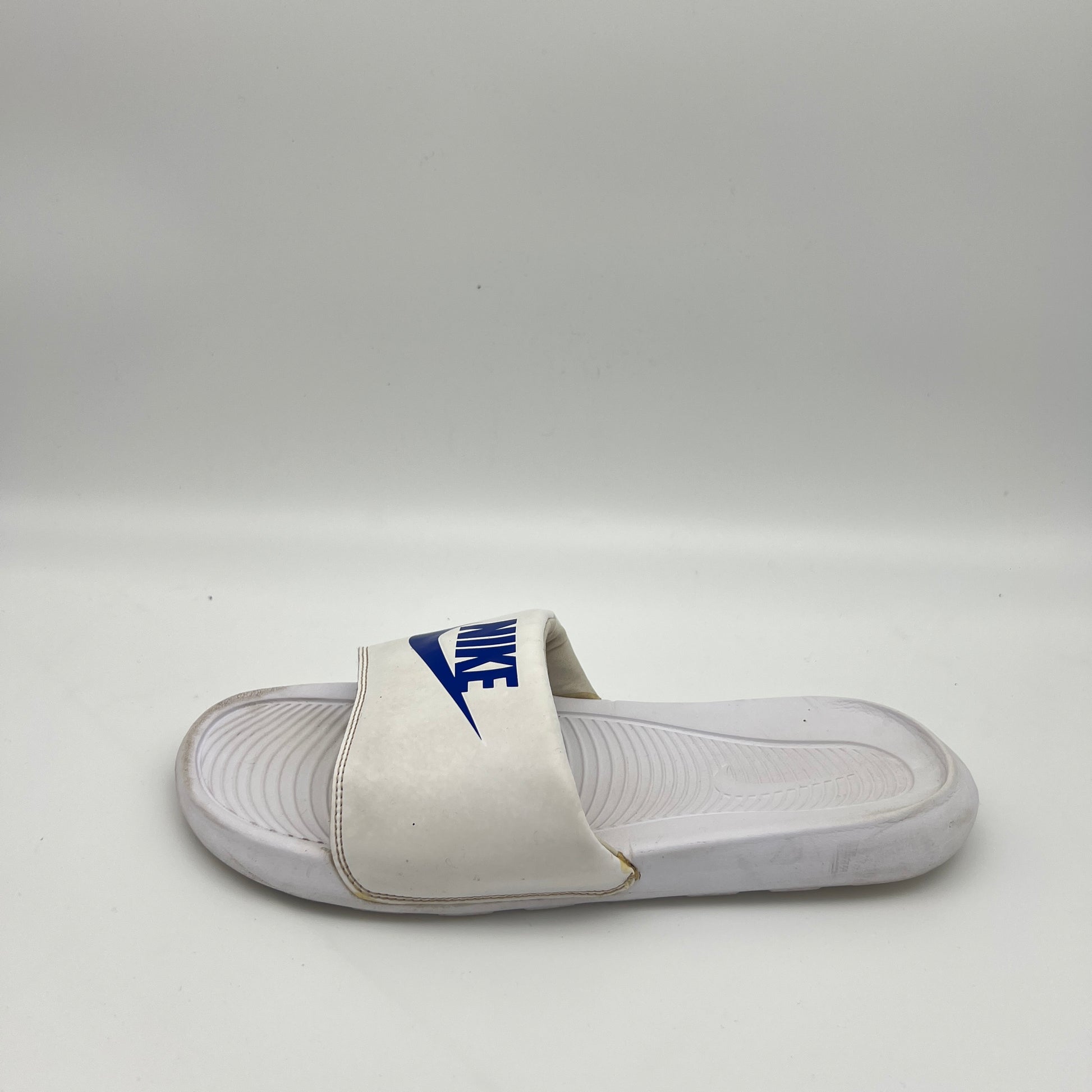 White Nike Victori One slide sandal with a royal blue Nike logo and swoosh on the padded strap, set against a white background.