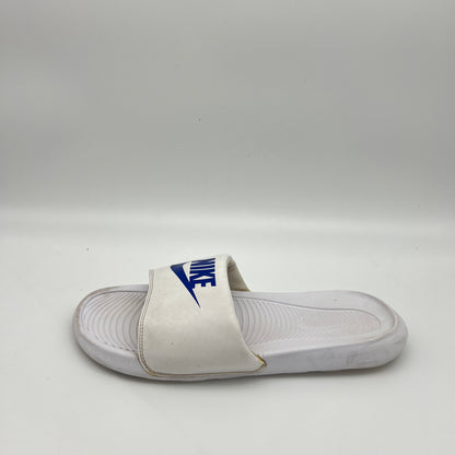 White Nike Victori One slide sandal with a royal blue Nike logo and swoosh on the padded strap, set against a white background.