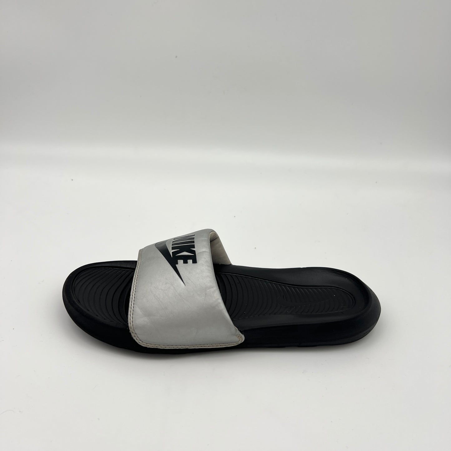 Metallic silver Nike Victori One slide sandal with a bold black Nike logo on the strap and a black contoured footbed.