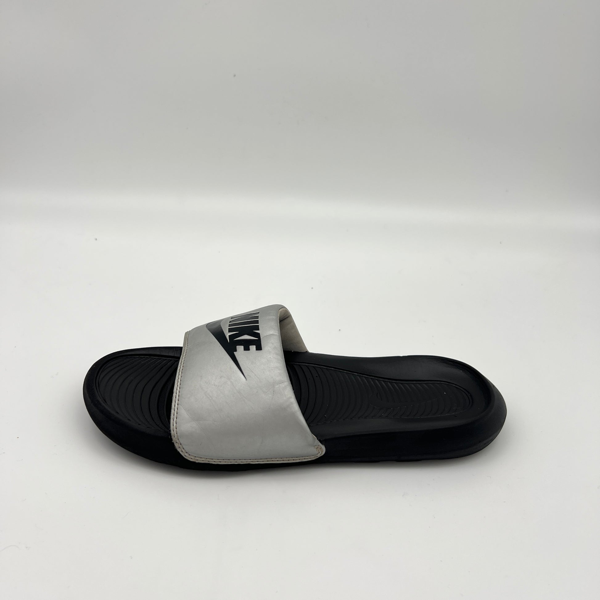 Metallic silver Nike Victori One slide sandal with a bold black Nike logo on the strap and a black contoured footbed.