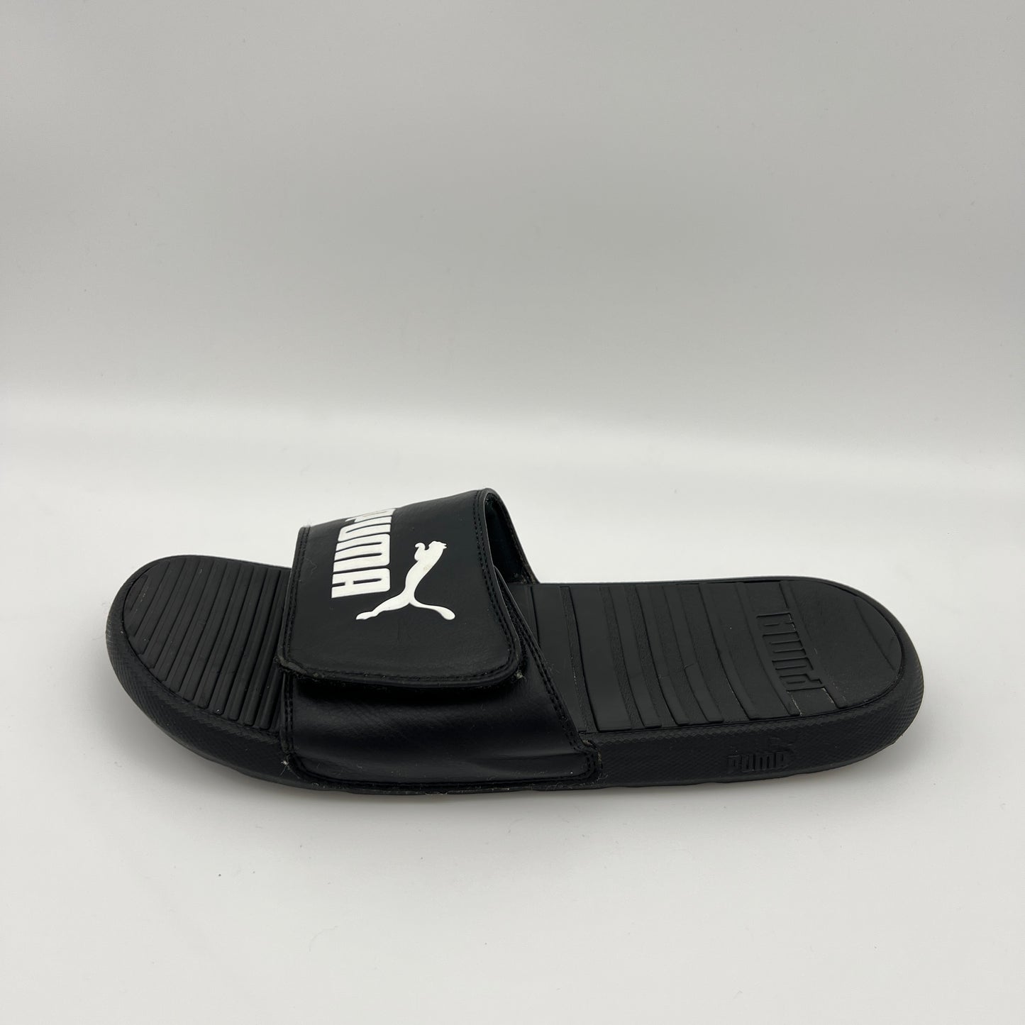 A side-angle view of a black Puma Cool Cat Sport slide sandal with a white Puma logo on an adjustable Velcro strap and a textured, contoured footbed.
