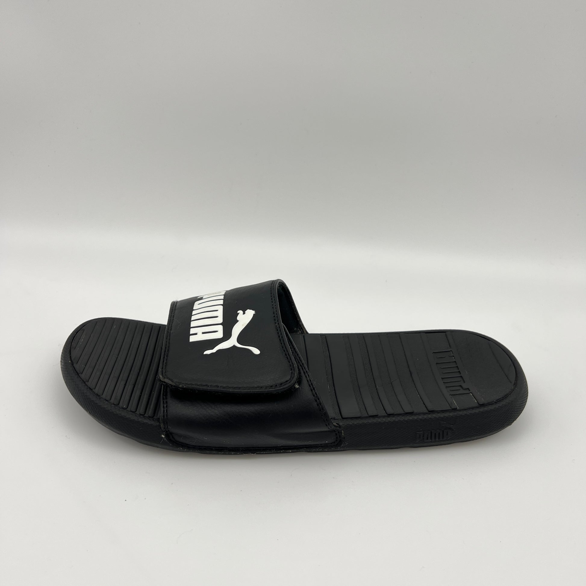 A side-angle view of a black Puma Cool Cat Sport slide sandal with a white Puma logo on an adjustable Velcro strap and a textured, contoured footbed.
