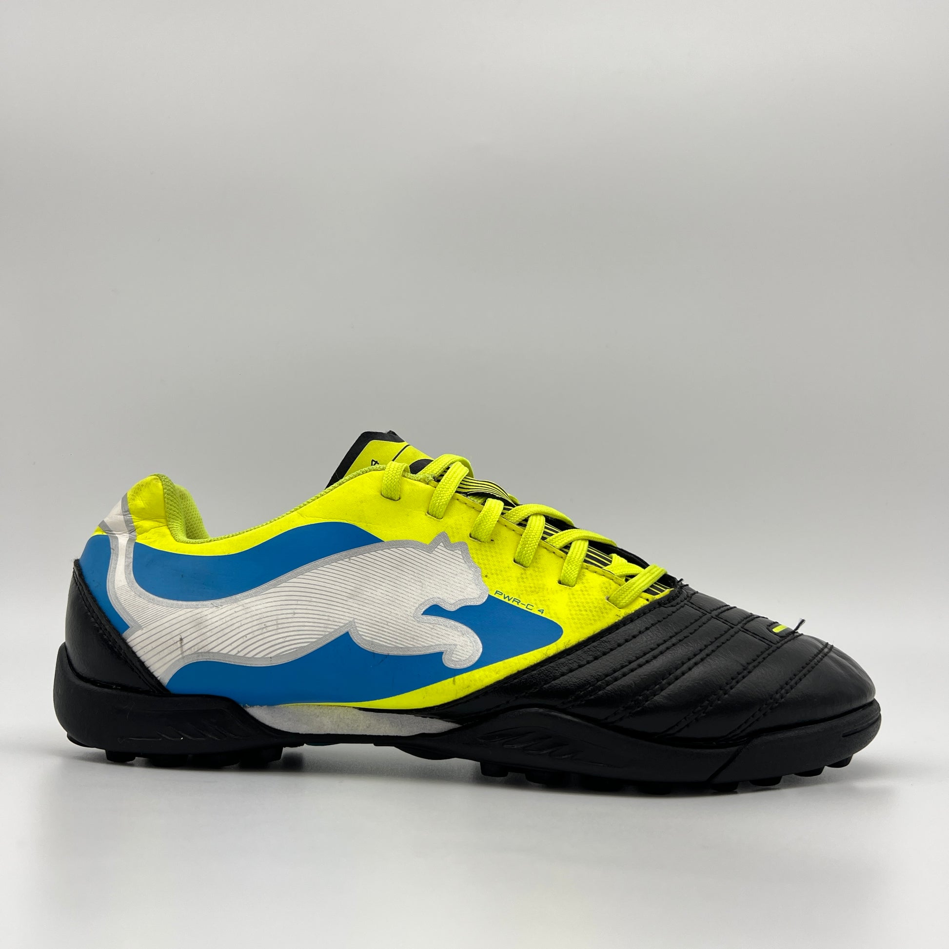 Side profile of a black and fluorescent yellow Puma PowerCat 4 TT soccer shoe with a large silver Puma cat logo over a blue graphic and a multi-studded black turf sole.