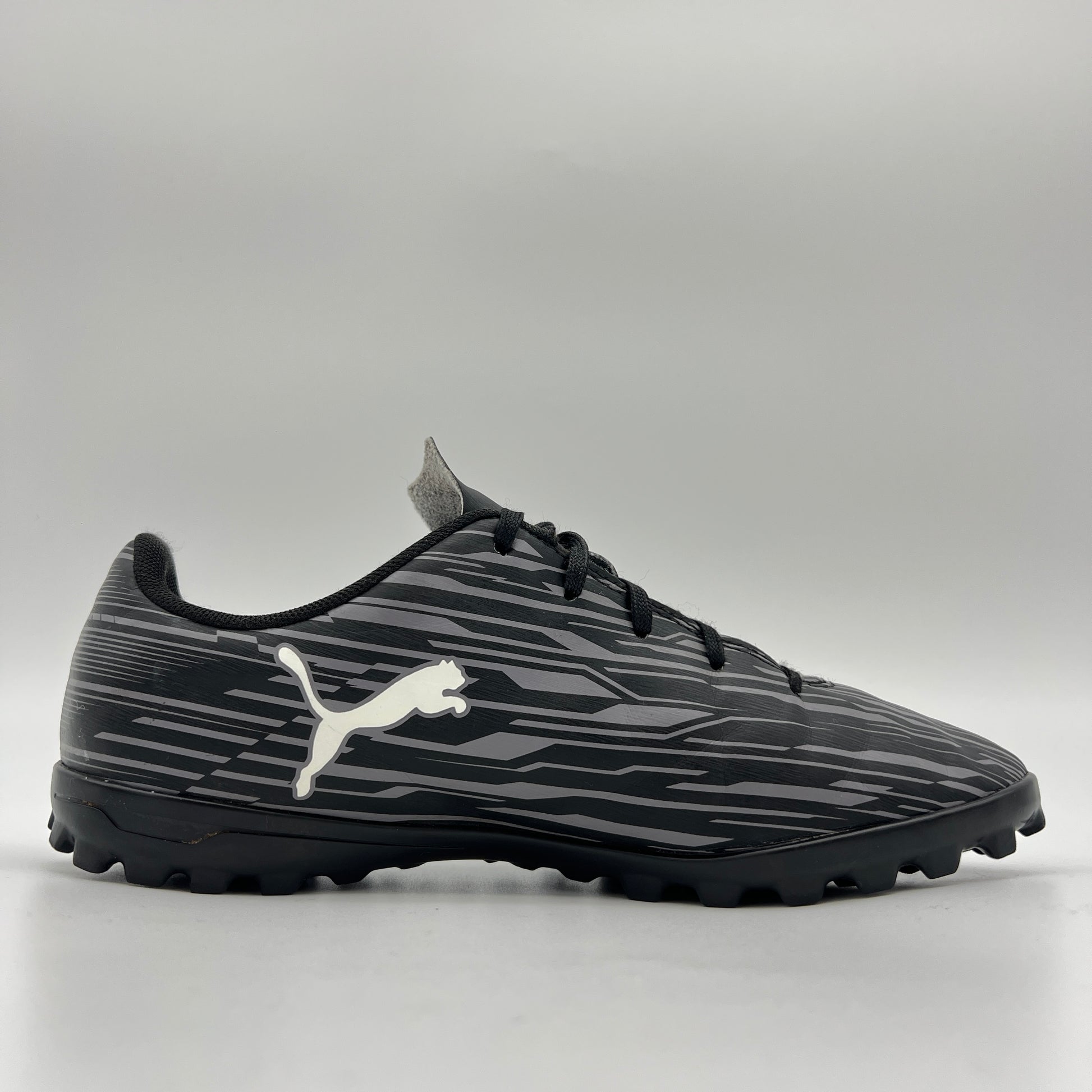 Side profile of a black Puma Rapido III TT soccer shoe featuring a grey-striped graphic pattern, white Puma formstrip, and black turf-ground studs.