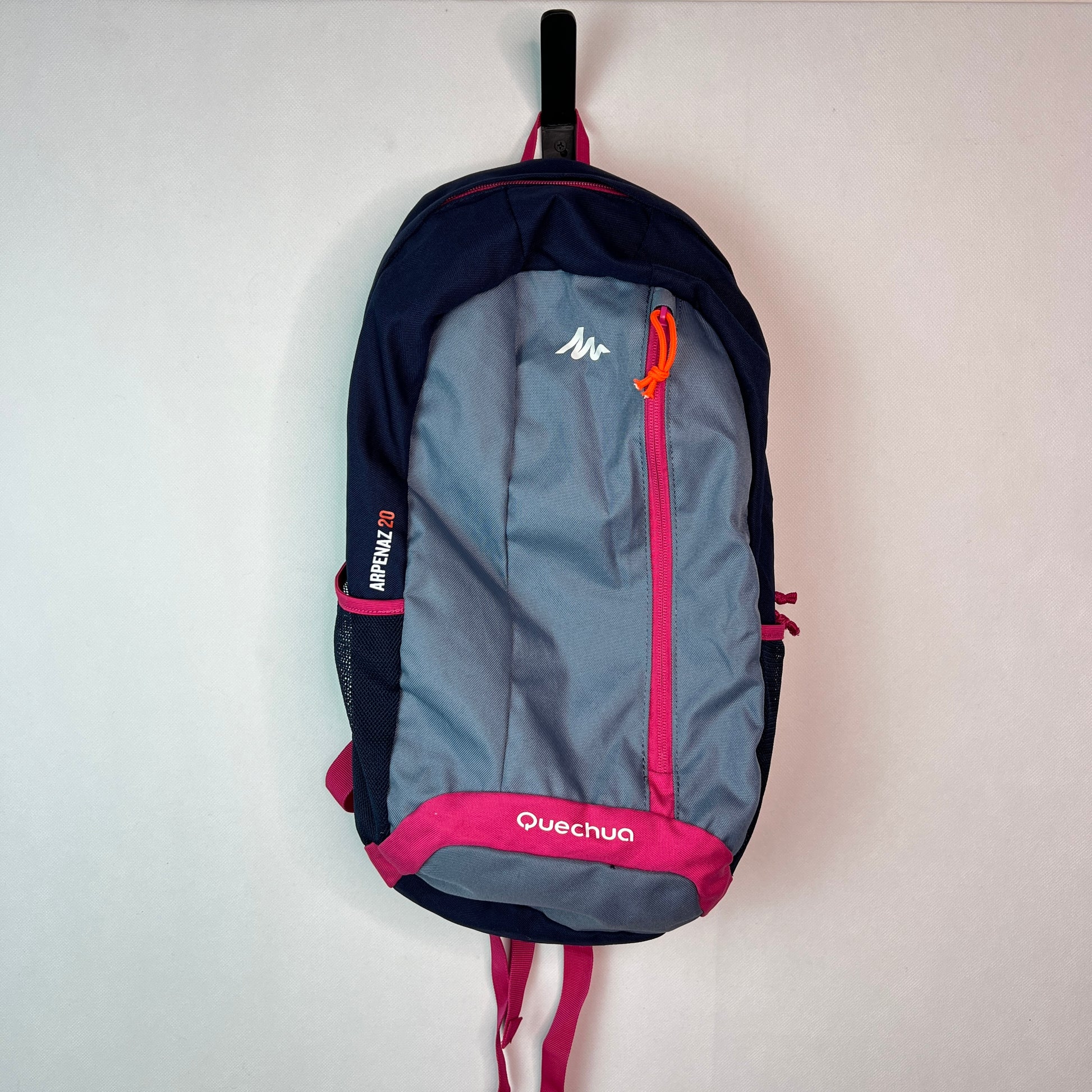 Front view of a grey and navy Quechua Arpenaz 20L backpack with pink accents and a vertical front zipper, hanging against a white wall