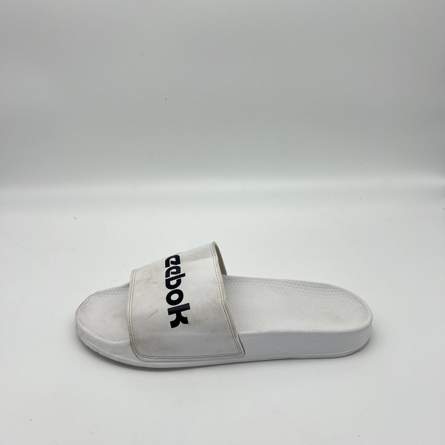 White Reebok Classic slide sandal with a bold black Reebok logo across the synthetic strap, featuring a textured white footbed.