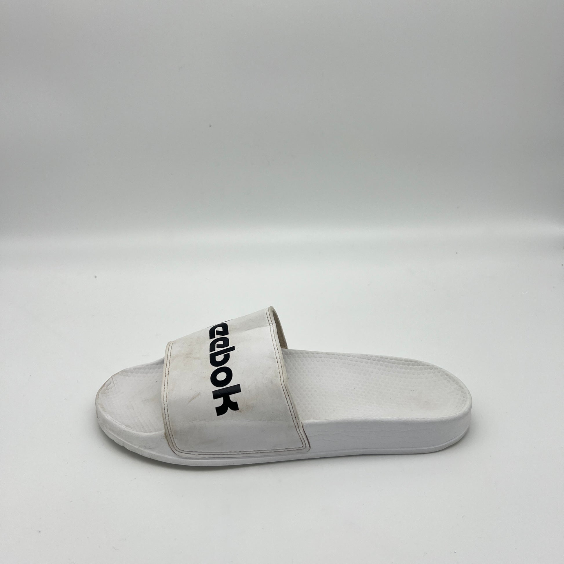 White Reebok Classic slide sandal with a bold black Reebok logo across the synthetic strap, featuring a textured white footbed.