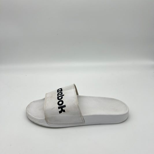 White Reebok Classic slide sandal with a bold black Reebok logo across the synthetic strap, featuring a textured white footbed.