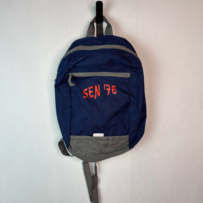 Front view of a navy blue SEN '76 backpack with grey trim and orange text, hanging from a black hook against a white wall.