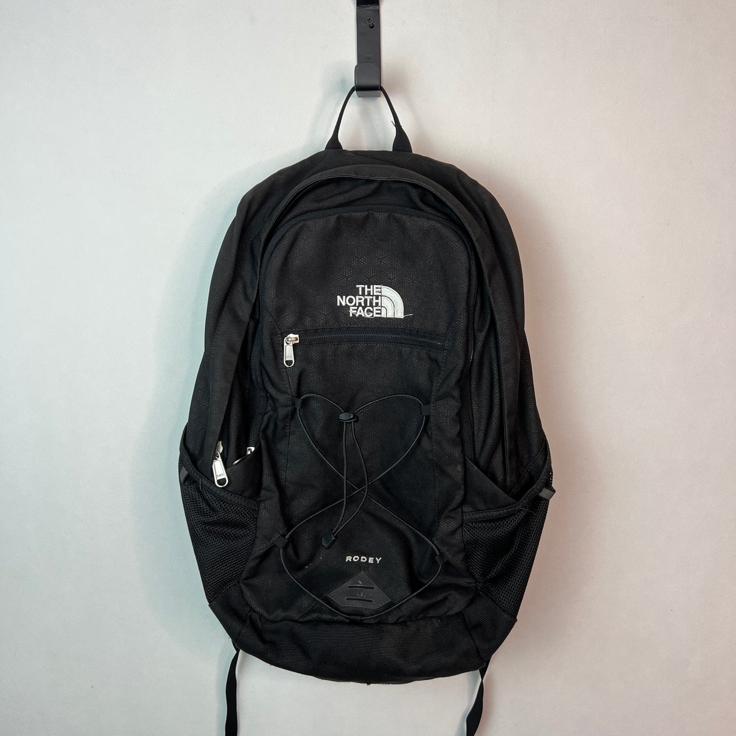 Black The North Face Rodey 27L backpack with front bungee system and white logo hanging on a white wall