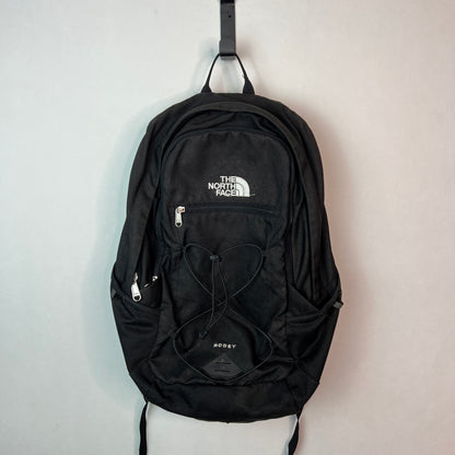 Black The North Face Rodey 27L backpack with front bungee system and white logo hanging on a white wall