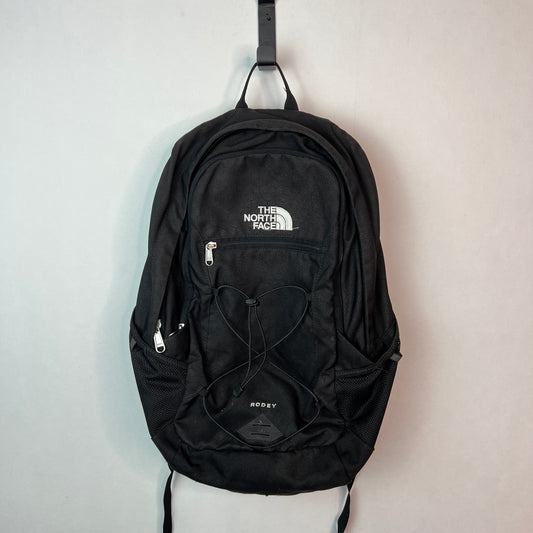 Black The North Face Rodey 27L backpack with front bungee system and white logo hanging on a white wall